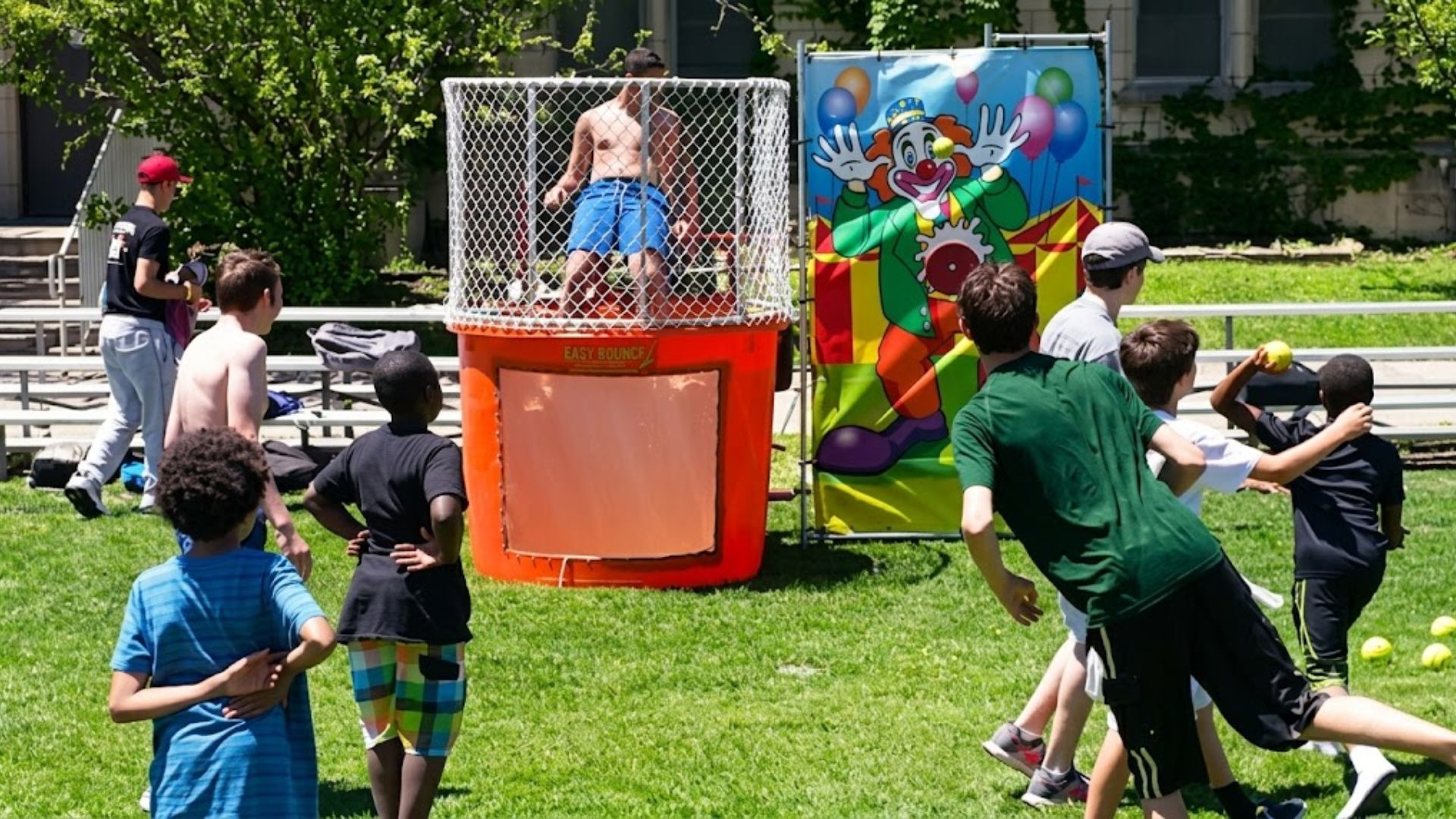 A teenager in swim trunks sitting on the seat of an orange dunk tank waiting to be dunked, next to a photo of a blue dunk tank ready for use at an outdoor festival